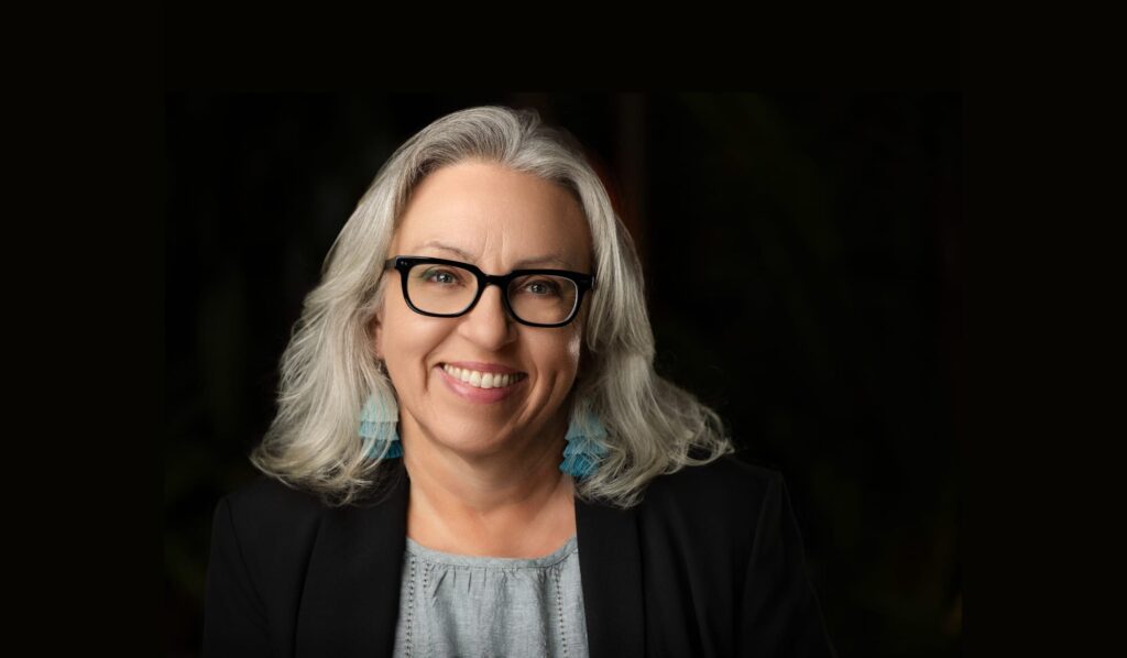 Portrait photo of Annelisa Stephan showing her smiling and wearing black glasses and a black blazer, with a blue-green blouse and blue-green tassel earrings