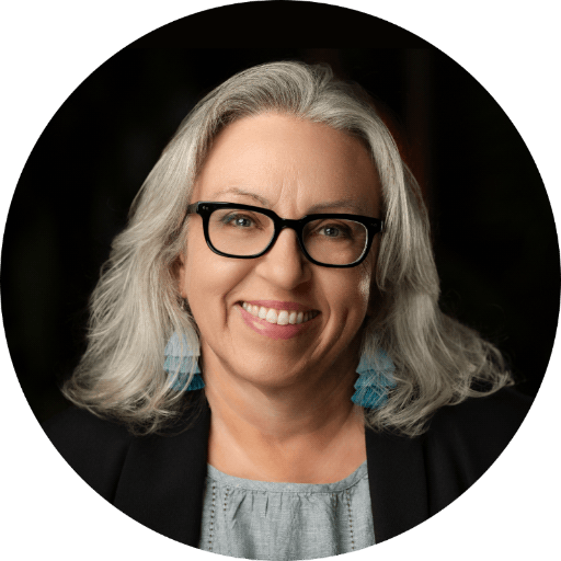 Portrait photo of Annelisa Stephan showing her smiling and wearing black glasses and a black blazer, with a blue-green blouse and blue-green tassel earrings